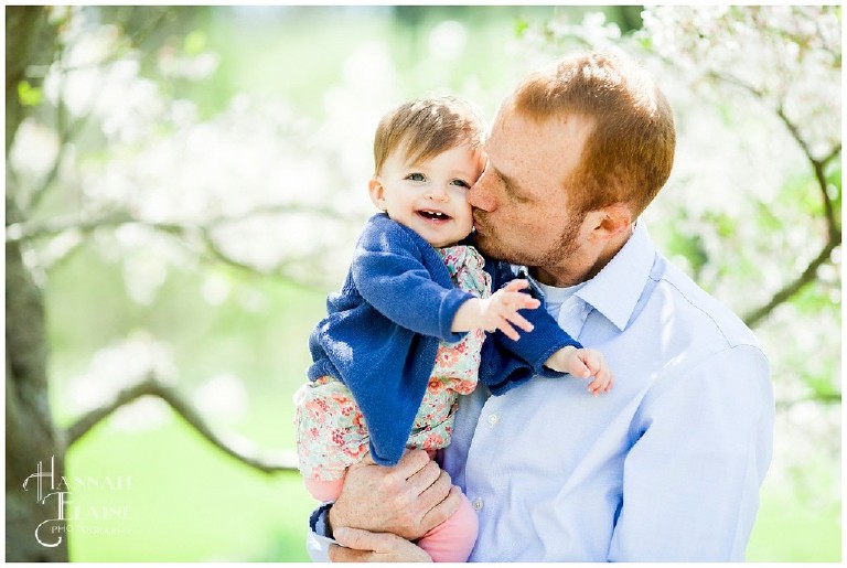 dad kisses daughter in the shade of a spring bloom tree
