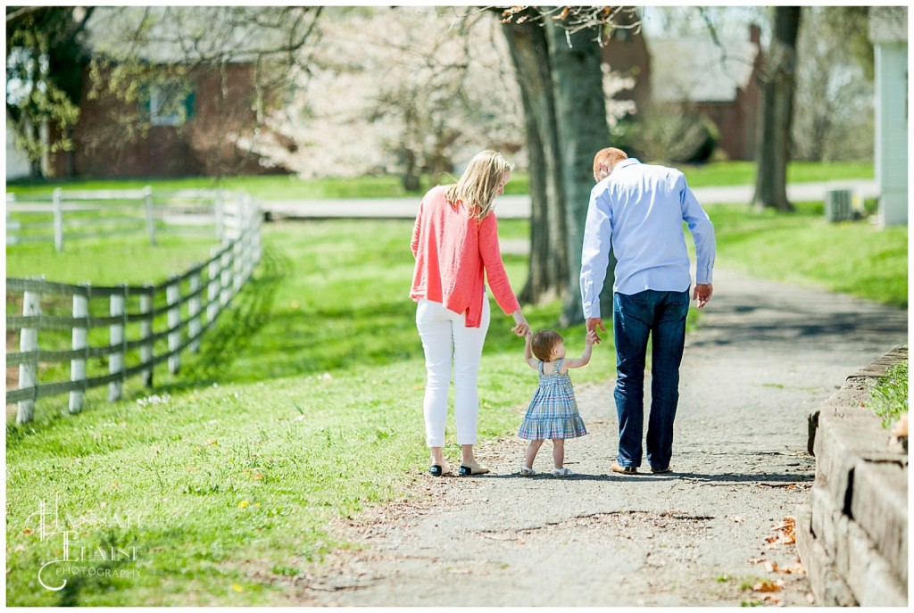 mom and dad help toddler on a walk down the path