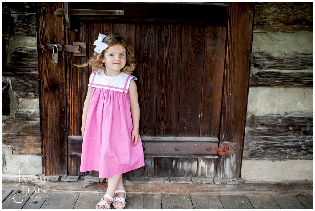 girl in pink smocked dress stands in front of rustic cabin door