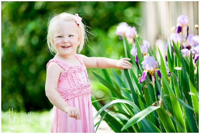 little blond girl smiles next to a garden of purple irises