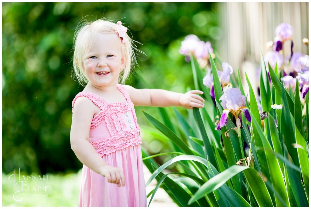 little blond girl smiles next to a garden of purple irises