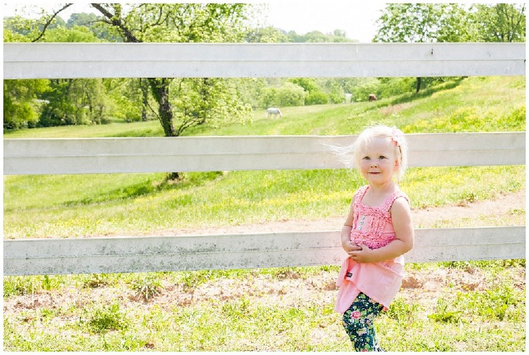 little blond girl stands against white fence with horses grazing in the distance