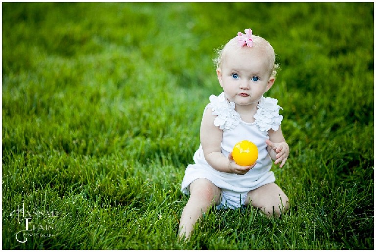 little blond girl sits in the grass holding a yellow ball