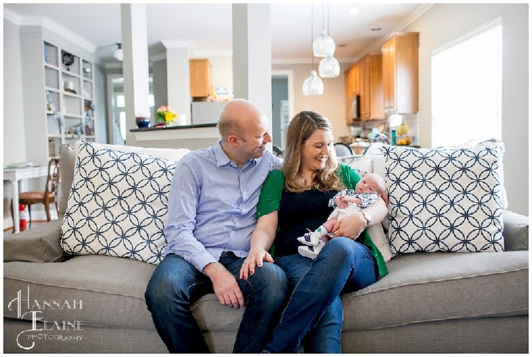 mom and dad smile at their baby son on their living room couch