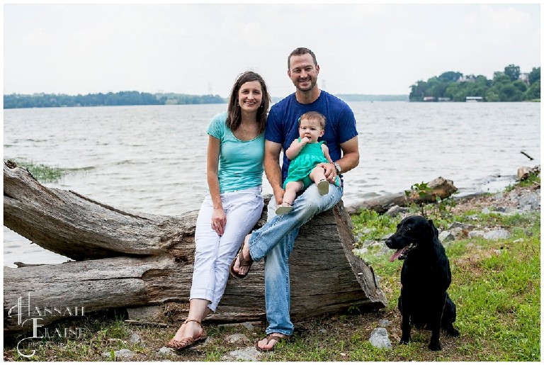 family of 3 sits with their dog by the lake