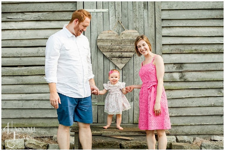 one year old girl learns to stand in front of rustic barn