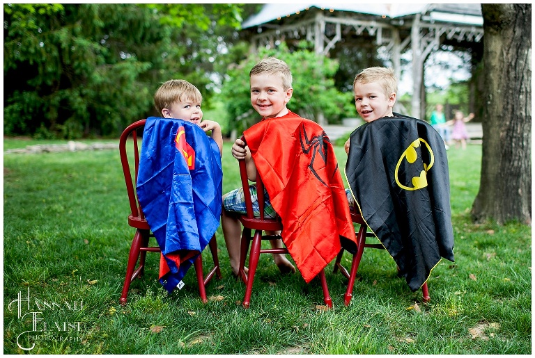 three brothers in super hero capes sit on vintage chairs