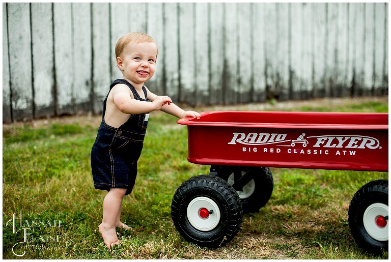 little boy in overalls holds onto radio flyer in front of antique barn